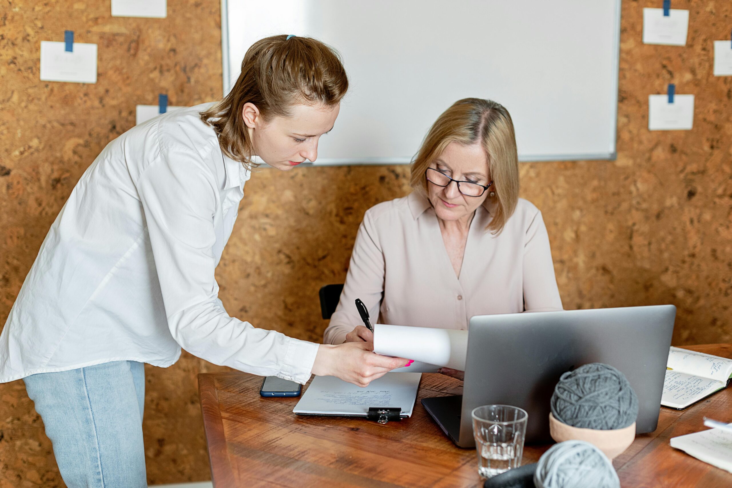 Two women discussing work documents in a modern office setting with a laptop.